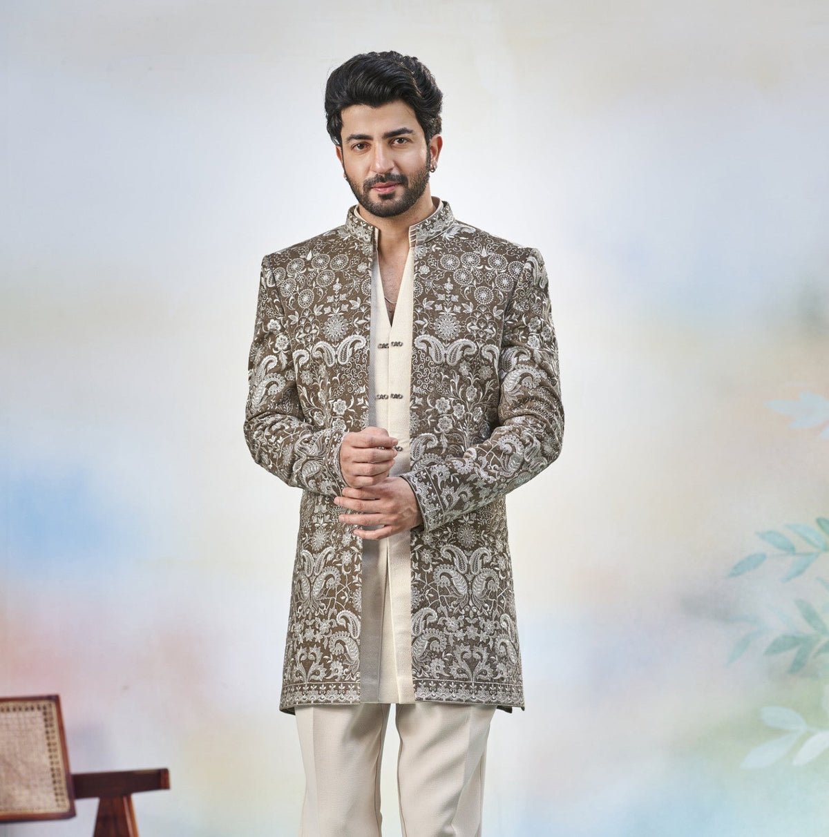 A man modeling a traditional Indian outfit featuring a brown and beige heavily embroidered sherwani jacket over a cream kurta and trousers, standing against a soft, light blue and white studio backdrop.
