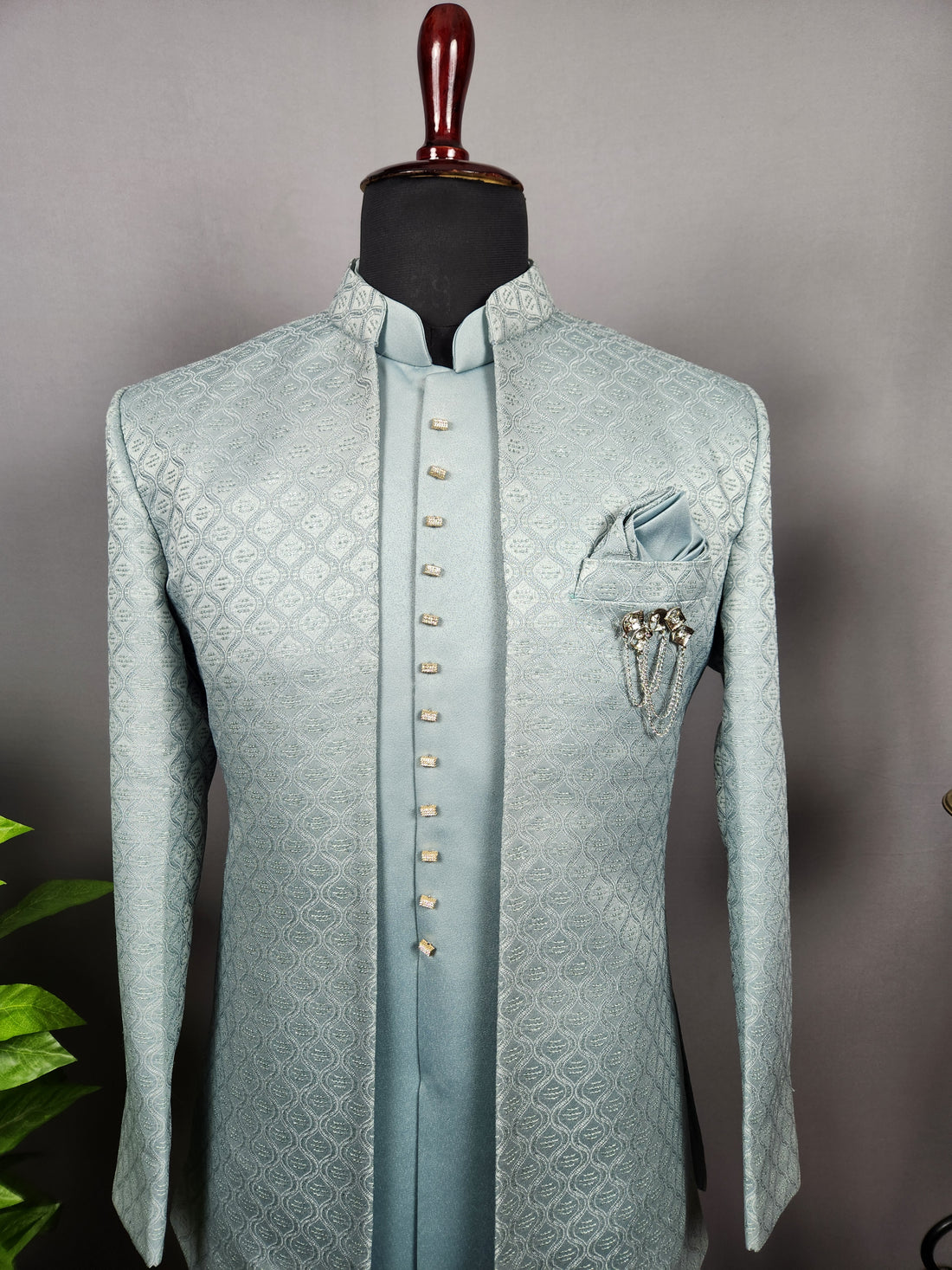 A man wearing a light blue embroidered sherwani and matching trousers stands in an ornate indoor courtyard with carved stone pillars and arches..