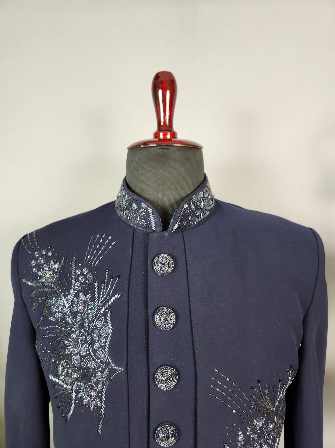 A man stands in a marble courtyard wearing a dark navy blue traditional embroidered sherwani jacket with matching trousers and brown dress shoes. A woman in a white lace dress sits in the background under an arched stone structure.