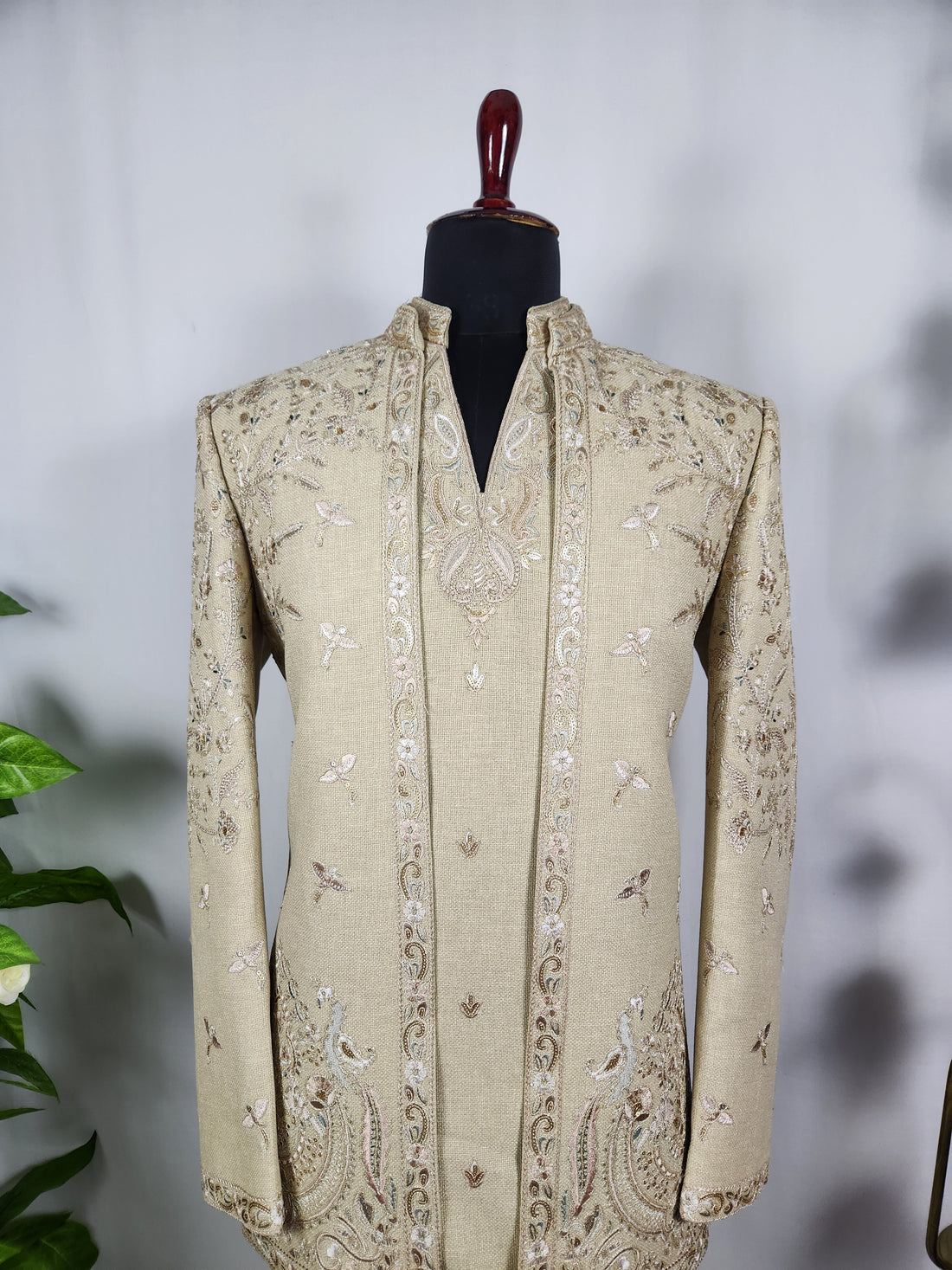 A man modeling a cream-colored embroidered sherwani jacket with matching trousers and dark brown dress shoes, standing on a stone balcony with a palace in the background..