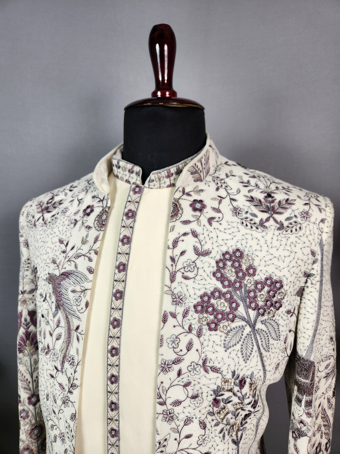 A man wearing a cream sherwani with purple floral embroidery and white trousers stands on a sunlit terrace by a lake, with a woman in a long white gown in the background..