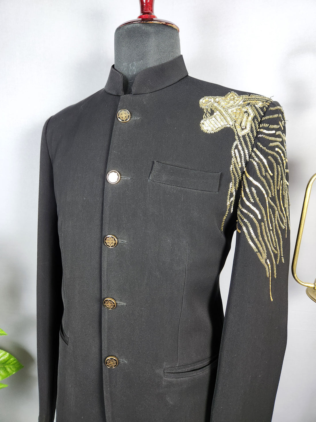A man wearing a black traditional Indian sherwani jacket with gold embroidery on the shoulder and gold buttons, paired with matching black trousers, standing in an ornate architectural corridor with arches and potted plants