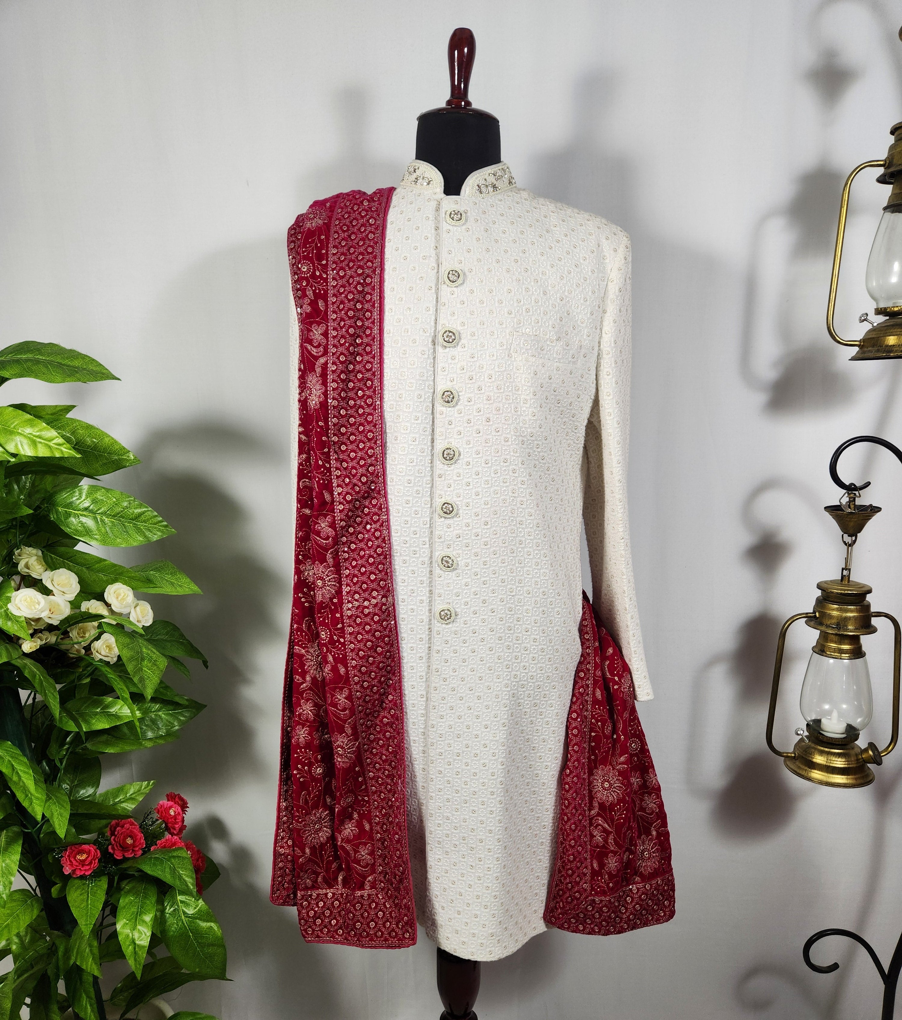 A white textured sherwani jacket with a red patterned dupatta draped over the shoulders, displayed on a dark wooden mannequin. The setup is styled with a green potted plant on the left and brass lanterns on the right against a white wall
