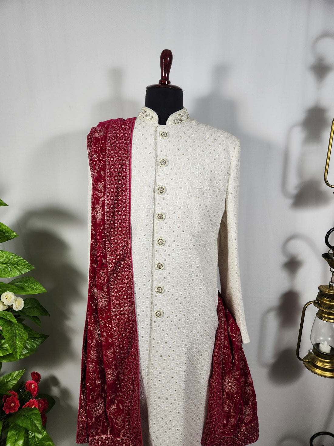 A white textured sherwani jacket with a red patterned dupatta draped over the shoulders, displayed on a dark wooden mannequin. The setup is styled with a green potted plant on the left and brass lanterns on the right against a white wall
